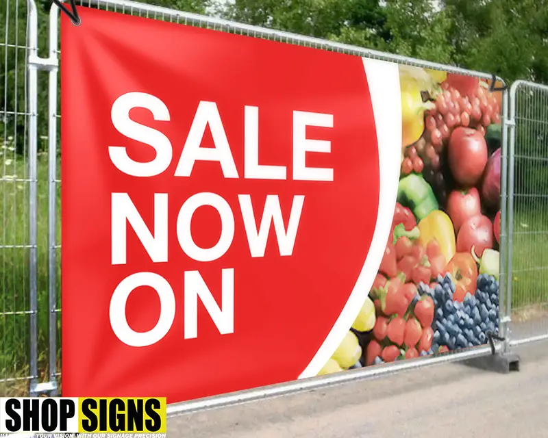 Outdoor PVC sale banner with bold red and white text and fruit graphics, displayed on a metal fence